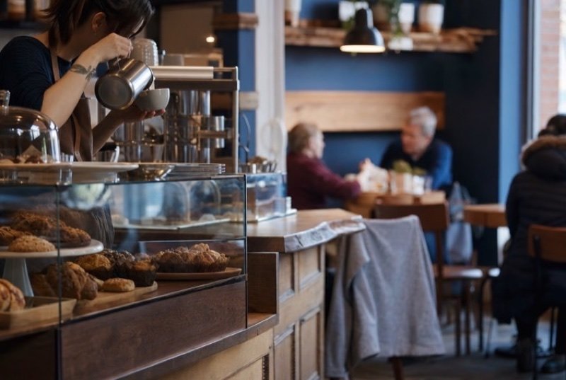 A café counter with baking under glass and a barista pouring milk, customers in winter coats behind