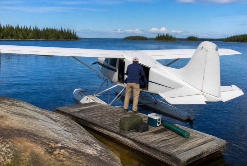 A float plane tied up at a wooden dock beside a granite shore, gear bags and a cooler on the planks