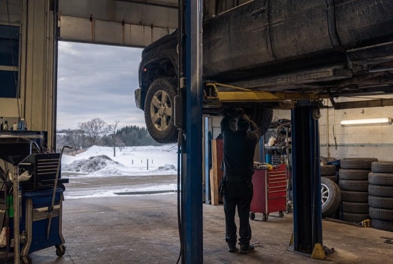 Mechanic under a lifted pickup in a small shop, a row of tires along one wall and snow visible through the bay door