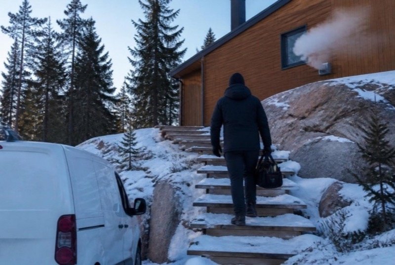 A heating technician carrying a tool bag up snowy steps to a wooden house built on pink granite